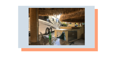 Photo of a Class A RV with a laptop on a table in the foreground taken from a chair under a tropical awning set outdoors